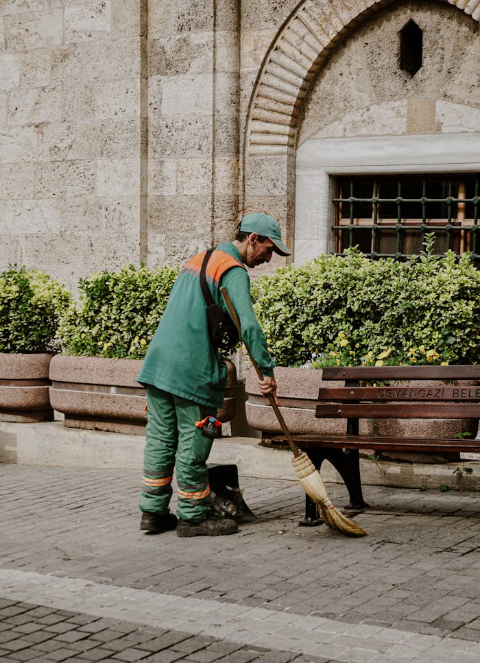 gallery-1 A street worker sweeps a park path in Turkey, wearing green uniform near leafy bushes.