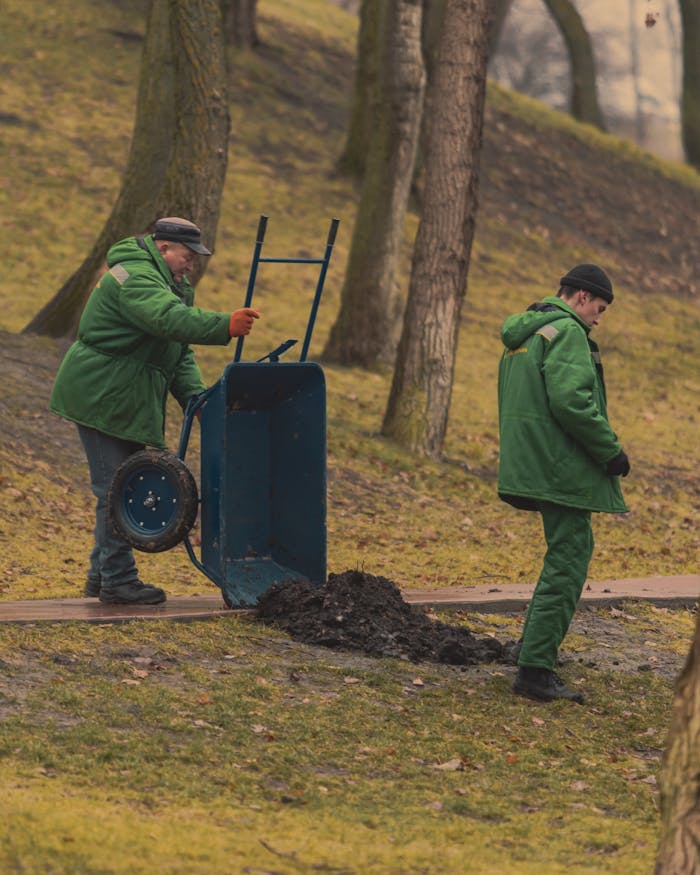 gallery-5 Two gardeners in green uniforms using a wheelbarrow in a park setting.