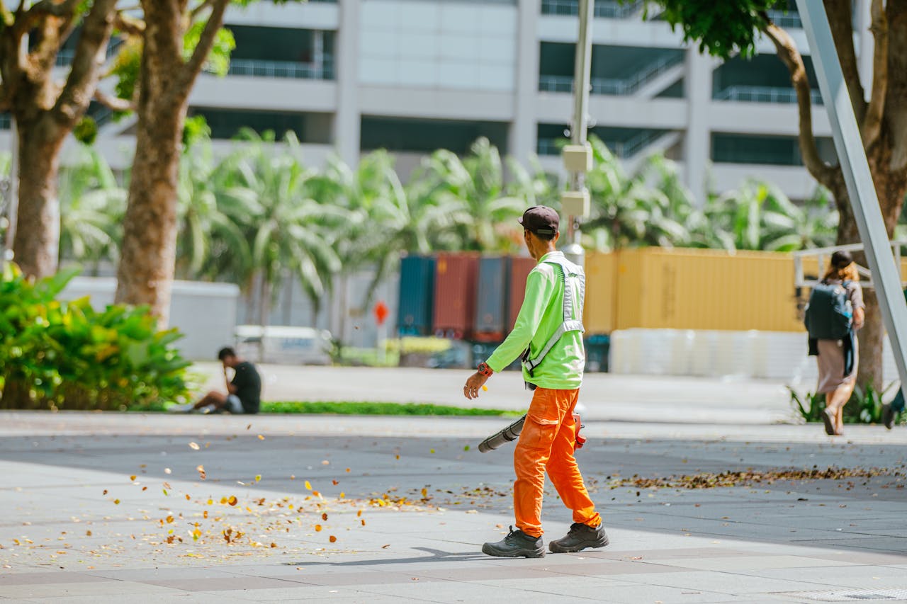 services-01 Street cleaner using a leaf blower in an urban park setting on a sunny day.