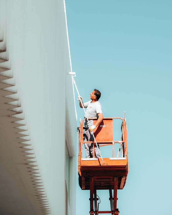 services-02 A man painting a building exterior from a cherry picker against a clear blue sky.