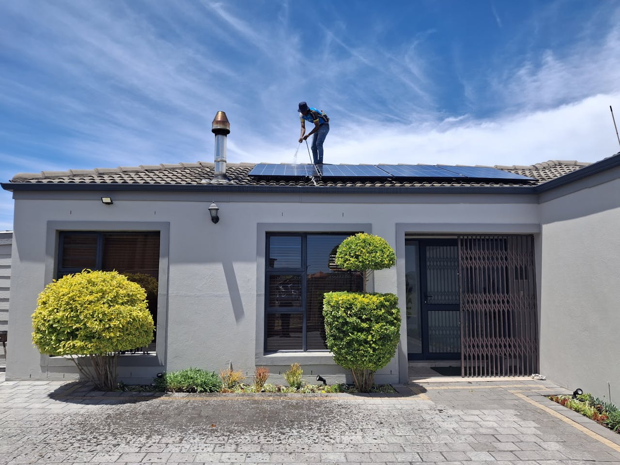 services-02 A person maintains solar panels on a house roof under a clear blue sky.