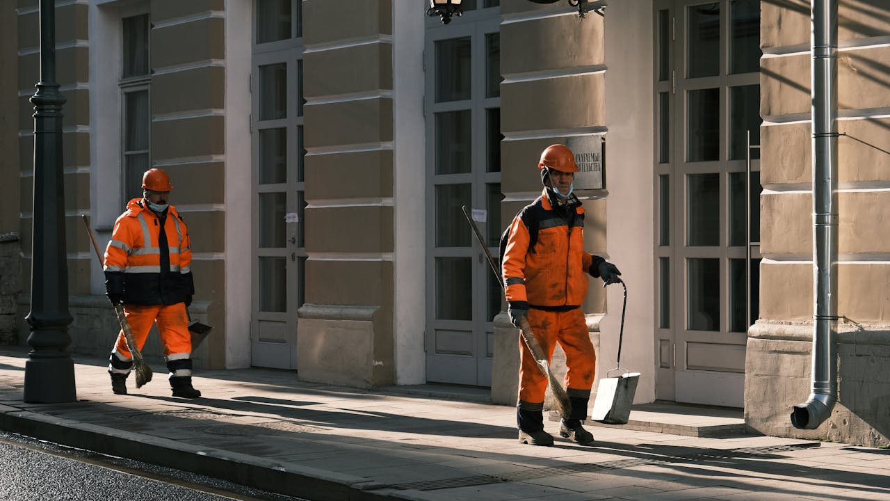 contact-img Street workers in orange safety uniforms walking along a city sidewalk in Moscow.